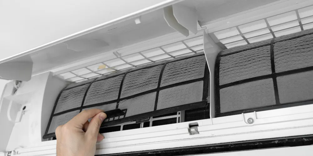 Close-up of a person's hand removing a dusty mesh filter from a white wall-mounted air conditioning unit for cleaning.