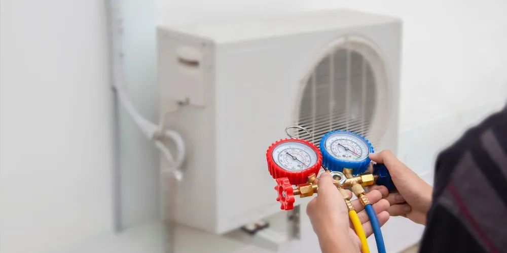 A technician holds a set of manifold gauges in the foreground, with yellow and blue hoses connected, while a white mini-split outdoor condensing unit is visible in the blurred background.