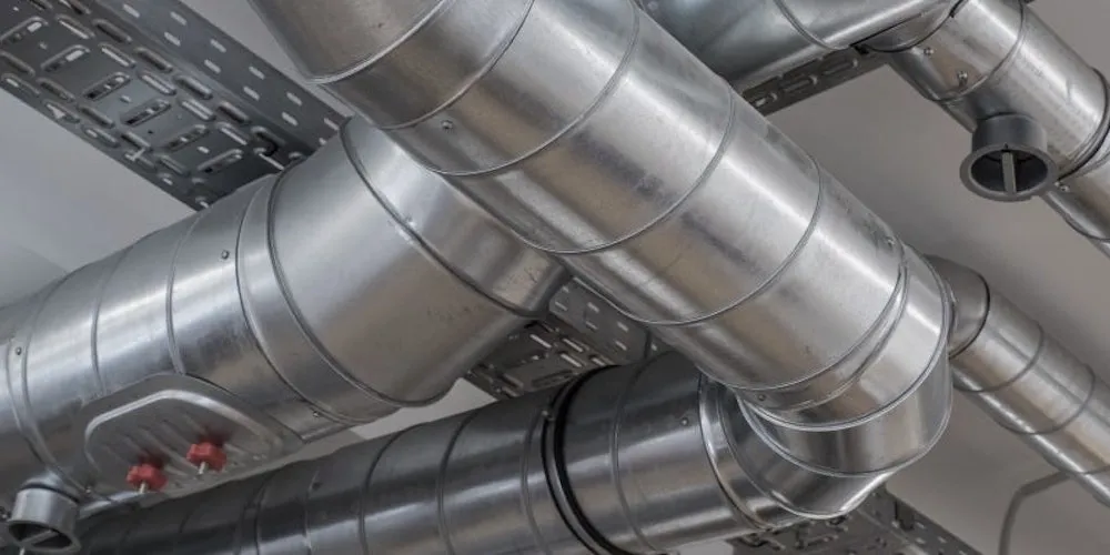 Close-up of multiple cylindrical spiral-seam galvanized metal ducts running parallel, with one T-junction and visible metal cable trays above them.