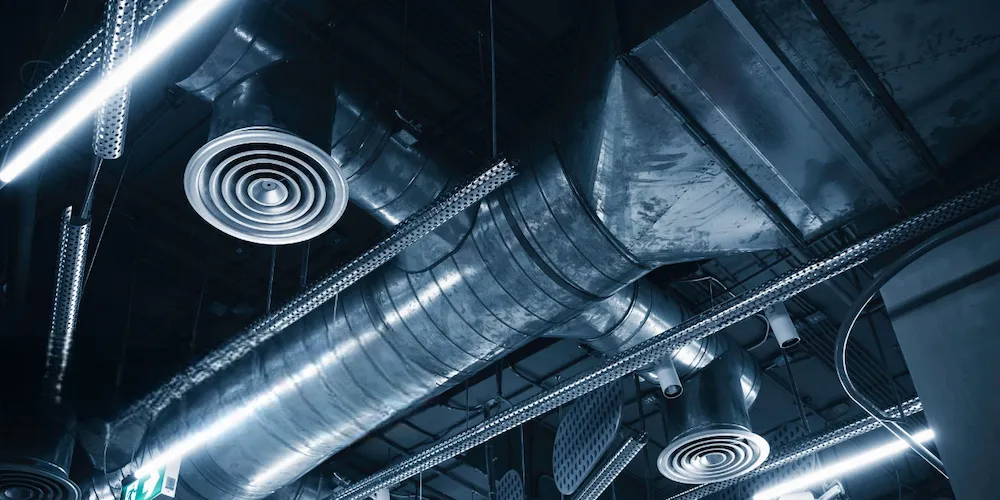 Low-angle shot of an industrial ceiling featuring exposed silver metal HVAC ductwork, circular air vents, and bright linear LED lighting.