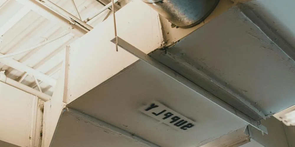 Close-up of large rectangular air ducts painted off-white in an industrial setting, showing hanging rod supports and a stamped identification number on the bottom surface.