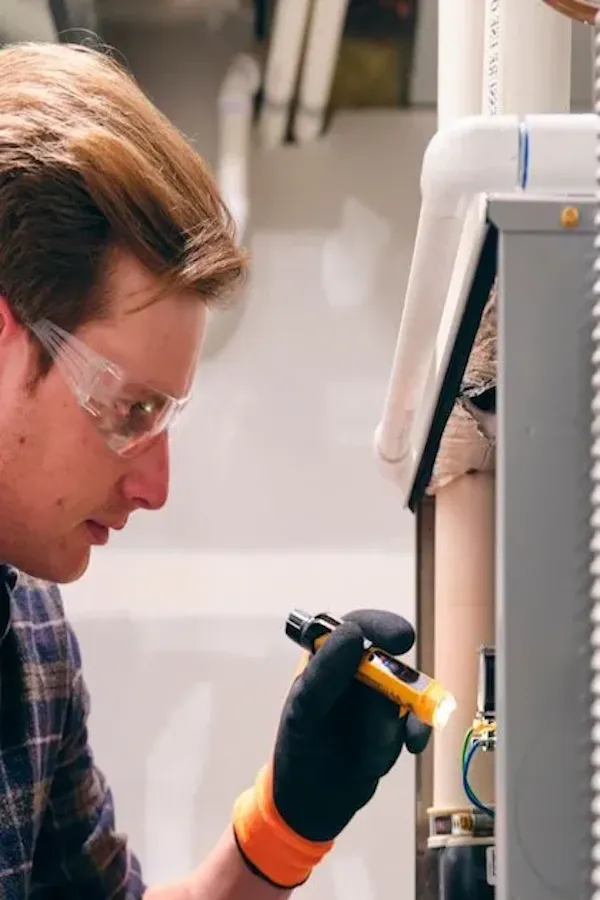A close-up profile of a male technician wearing safety glasses and black work gloves. He is using a small yellow flashlight to carefully inspect the internal wiring and components of an open gray electrical or HVAC panel.